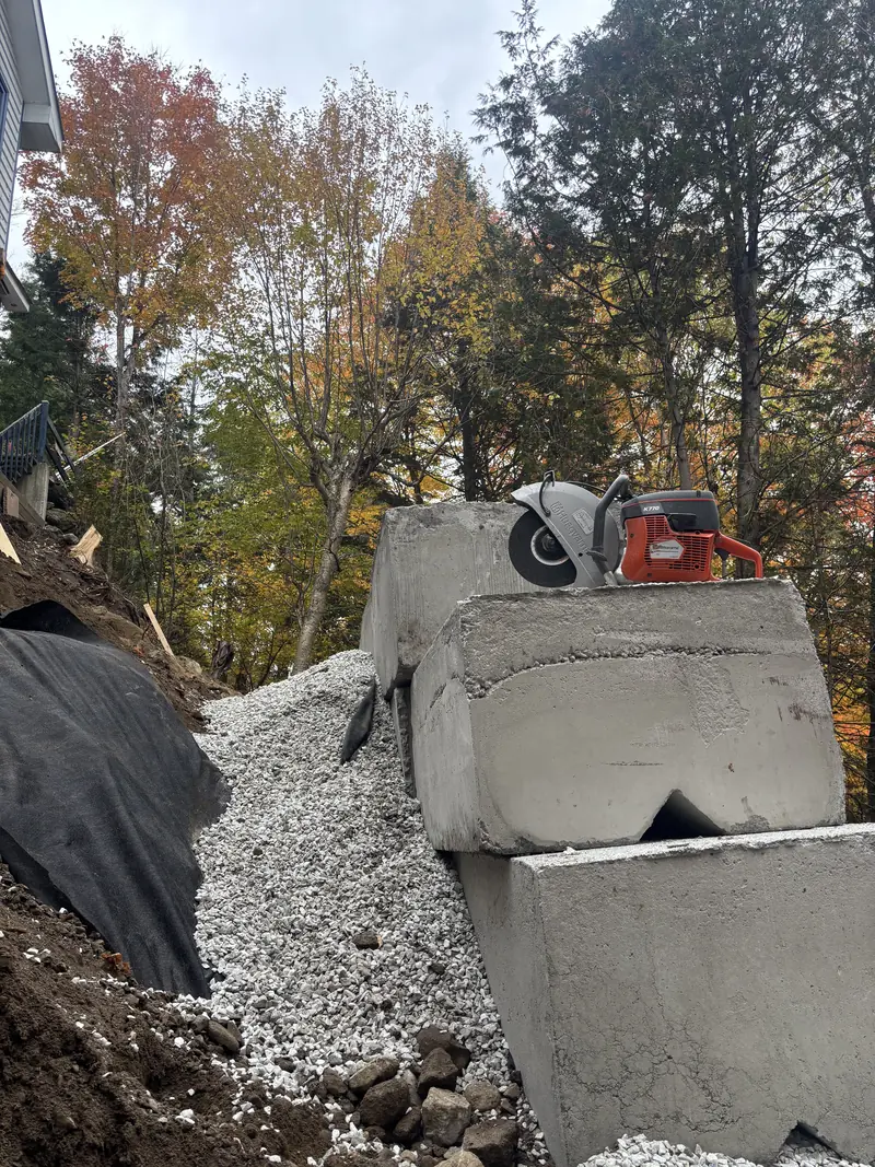 Pose de gros blocs de béton préfabriqués pour un mur de soutènement, avec remblai de pierre concassée et membrane géotextile sur la pente Pose de gros blocs de béton préfabriqués pour un mur de soutènement, avec remblai de pierre concassée et membrane géotextile sur la pente