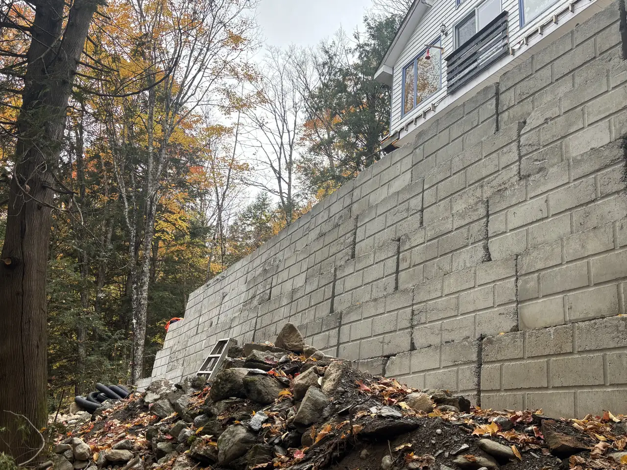 Haut mur de soutènement en grands blocs de béton gris soutenant une maison sur un terrain boisé en pente Haut mur de soutènement en grands blocs de béton gris soutenant une maison sur un terrain boisé en pente